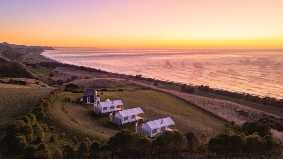 Sunset over the Kekerengu coast, a row of canvas tents perched on the hill above a long, empty beach, Kekerengu surf spot, Ka