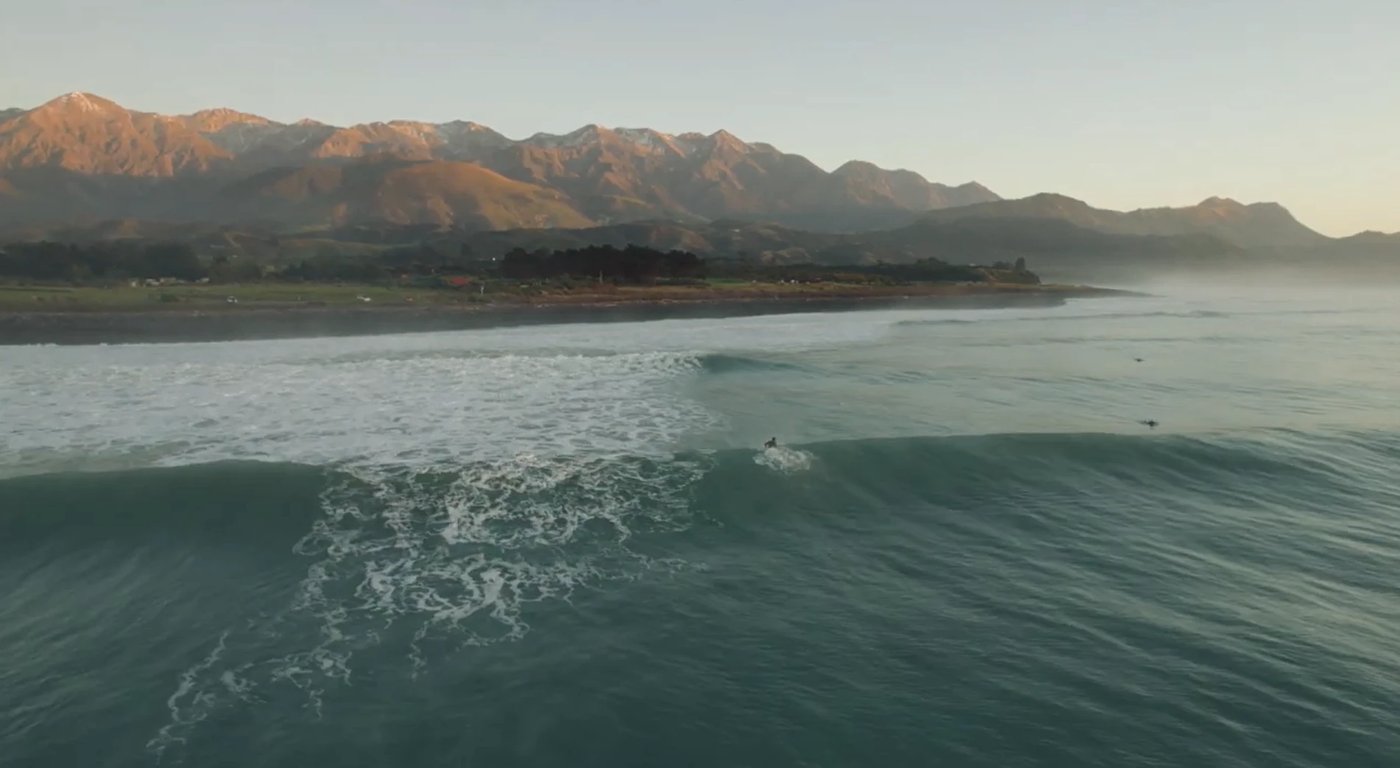 First light catching the snow on the Kaikōura ranges as a surfer drops into a clean dawn wall, Meatworks surf spot, Kaikoura 