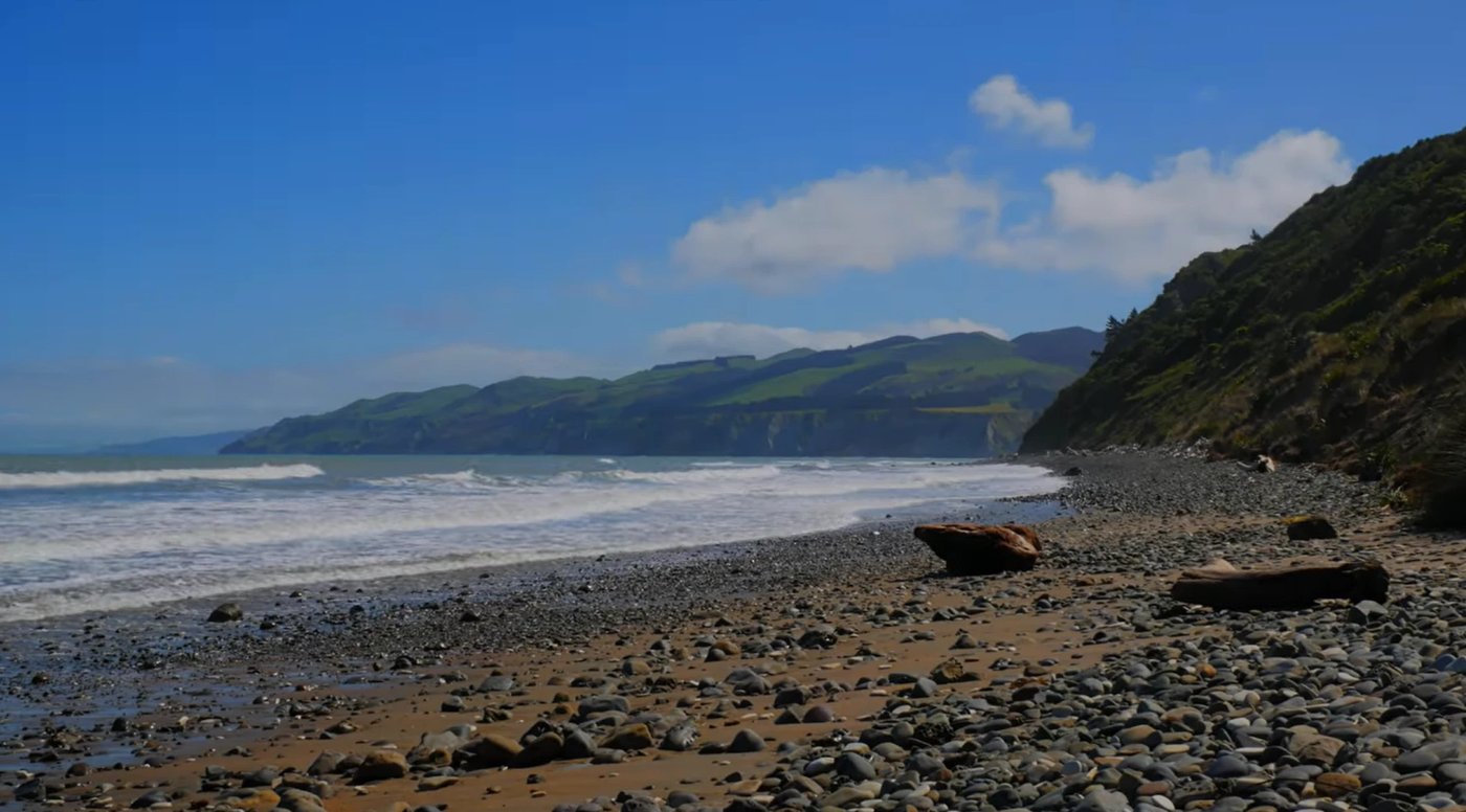 Driftwood and cobbles line the quiet shore, the green North Canterbury hills rolling away down the coast, Motunau Beach surf 